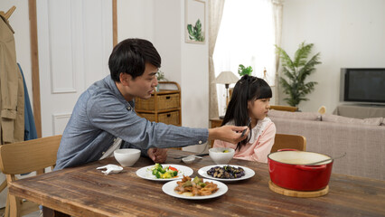 asian girl picky eater turning head and saying no to her dad as he is encouraging her to take a bite of vegetable. he holds the food with chopsticks near her - obrazy, fototapety, plakaty