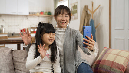 smiling asian mother and daughter waving hi to phone screen while making video call to friends at home. the girl sitting near her mom and saying greetings