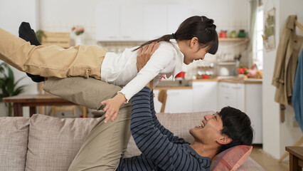 Side view happy Asian father lying on sofa and holding up daughter on his legs at home. The girl open arms and flying in air like plane