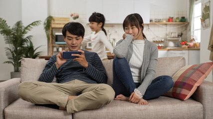 bored asian mother sitting on sofa looking at energetic daughter running around in living room while the addicted father is playing with mobile phone and ignoring her