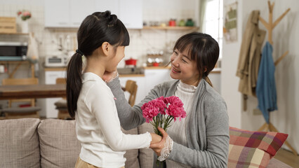 asian girl walking near to show surprise carnation bouquet to mother on mother's day at home. the happy mom smelling the flowers and smiling at her daughter while touching her face