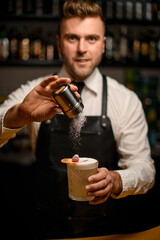 hand of barman holds glass with foamy cocktail and sprinkle it with powder
