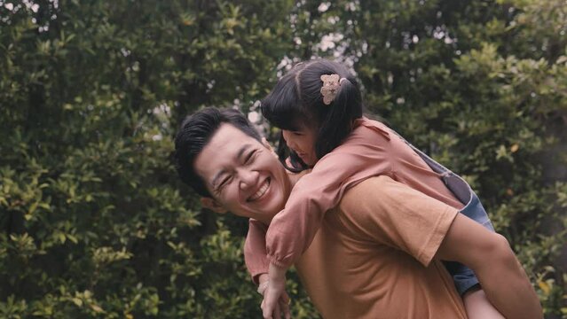Young father giving his daughter piggyback and smiling in the outdoor park. Happy Asian parent.