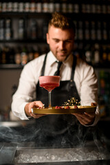 hand of barman holds round tray with raspberries and flowers and glass of cocktail on it