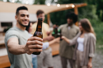 Happy young Arab guy with diverse friends toasting with beer bottles, drinking alcohol near RV outdoors, selective focus