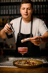 male hand holds stick with marshmallows over glass of cocktail and sets it on fire with gas burner