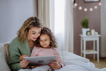 Happy mother with her little daughter lying on bed and using tablet at home. - Powered by Adobe