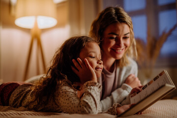 Happy mother with her little daughter lying on bed and reading book in evening at home.