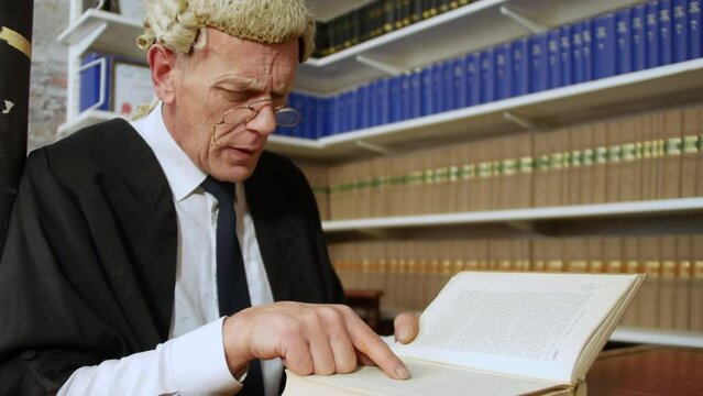 Close up of a judge or barrister reading a law book in the Judge's chambers library. The man is wearing a wig and gown with glasses.