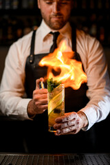 hand of man holds cocktail glass with cucumber slices and mint, sprinkles and carefully sets it on fire