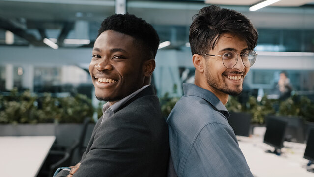 Portrait two multinational male colleagues men African man arab Indian manager posing in office company standing back to each other partnership looking at camera smiling business partners employees