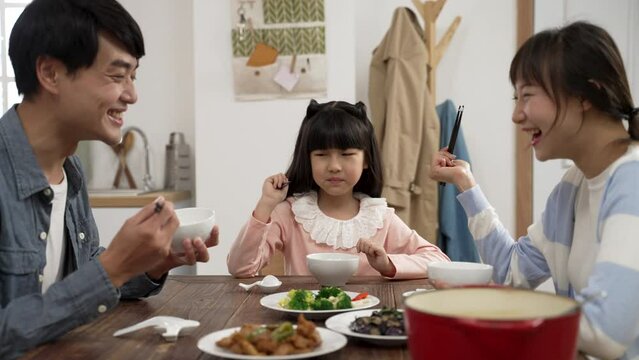 happy asian family of three talking and laughing while having lunch together at home. the dad helps pass the meat dish to her daughter and she accidentally drops the food
