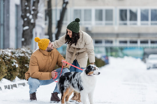 full length of cheerful young couple in winter jackets looking at each other near australian shepherd dog.