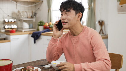 asian man picking up call while eating lunch alone at home. he puts down chopsticks and ball to...