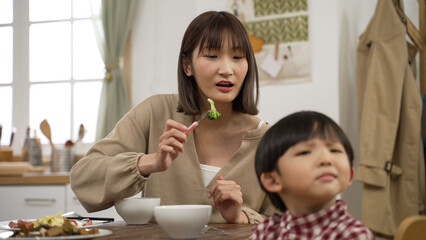 selective focus frustrated Asian mother holding a fork with vegetable and asking his baby son to...