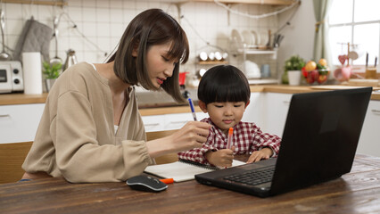 Asian mother supporting her son to write schoolwork at dining table while he is learning online from home with a laptop computer