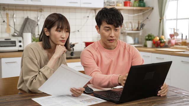 Frustrated Asian Couple Managing Finances For The Tax Season With Laptop In The Dining Room At Home. They Prop Head Feeling Worried About Financial Problems While Looking At Document From Bank