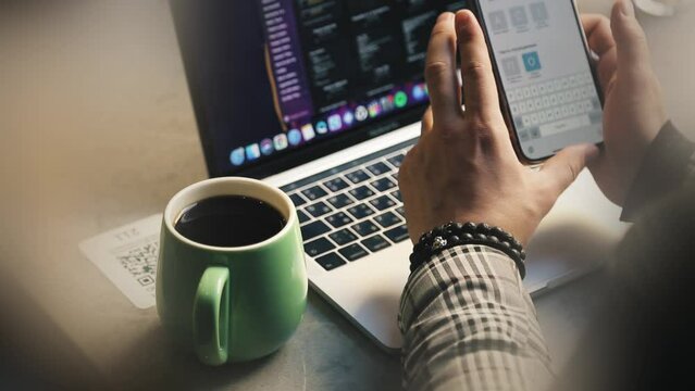 A man sits working at a computer, drinking coffee and holding a phone in his hands. Workflow, comfort for a freelancer. Male hands close up.