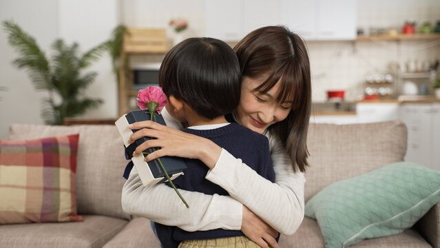 Asian baby boy show his mother the carnation flower and Mother&rsquo;s Day gift. smiling mother giving her son a big hug in living room
