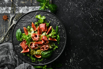 Green salad with prosciutto, cucumber and fig on a black stone plate. Food. Top view. Free space...