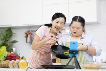 mother and down syndrome teenage girl or her daughter cooking food together and live streaming online via smartphone on tripod in a kitchen