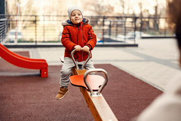 A happy three year old boy rocking on seesaw at playground with his mother of nanny/babysitter.