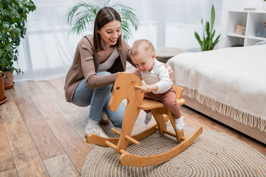 Young Woman Holding Baby On Rocking Horse At Home.