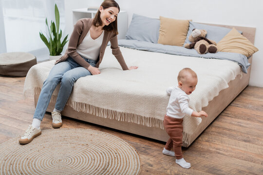 Positive woman sitting on bed near baby son walking in bedroom.