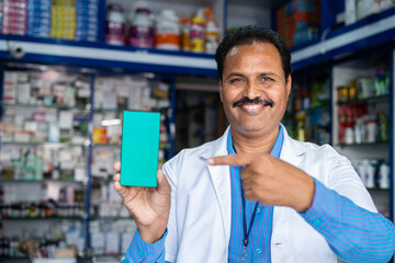 Smiling medical store pharmacist in uniform showing green screen mobile phone by pointing finger at...