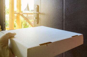 Close-up of a cardboard pizza box, a courier's hand in a rubber glove, holding a food delivery box against the background of an open door in a country house