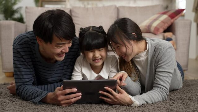 happy Asian family of three watching funny online video with tablet computer together at home. they lie prone on living room floor and point at screen while laughing