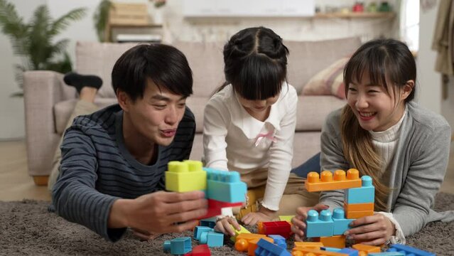 slow motion of happy asian dad, mom and daughter playing building blocks together on floor in the living room at home. childlike parents having fun making their toy collide