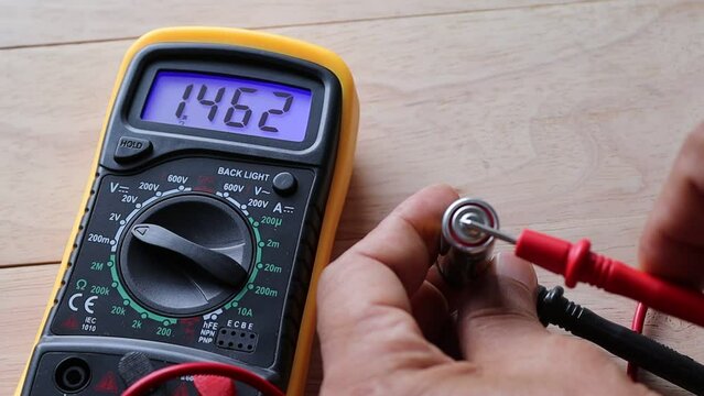 Hands are using a digital multimeter to measure electric voltage for checking battery cell 1.5 VDC on wooden background closeup. 