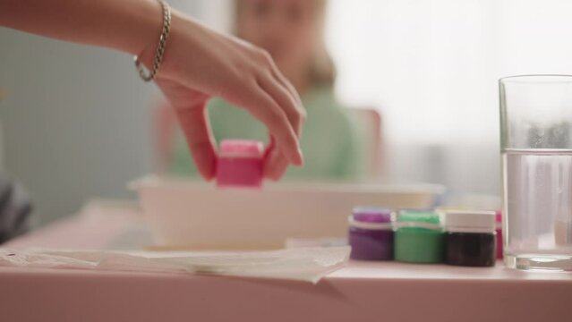 Teacher takes bottle with pink paint while little girl sits at table in classroom slow motion. Preparation for Ebru drawing lesson. Supplies for artistic work