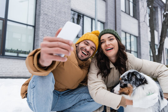smiling man in knitted hat taking selfie with girlfriend and australian shepherd dog in winter.