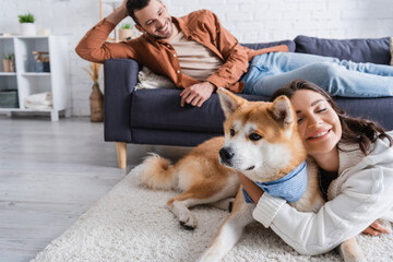 cheerful young woman hugging akita inu dog near boyfriend on couch.