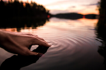 Closeup of a hand touching the water making smooth waves on the surface in a lake in sunset - obrazy, fototapety, plakaty