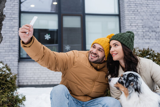 happy man in knitted hat taking selfie with girlfriend and australian shepherd dog in winter.