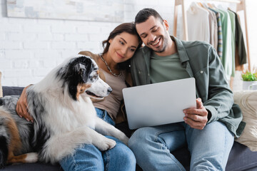 positive young couple watching movie on laptop near australian shepherd dog.