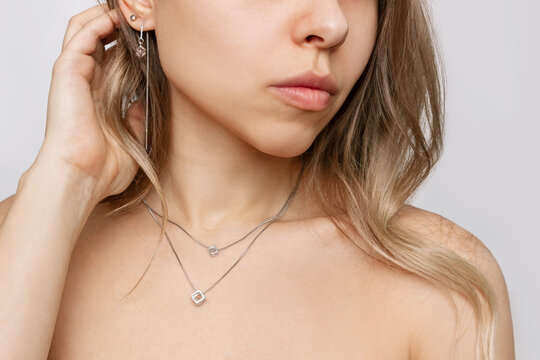 Cropped shot of young caucasian blonde woman with wavy hair wearing elegant diamond earrings and silver chain with a pendant isolated on a gray background. Jewelry with gemstones, holiday accessories