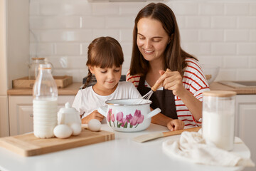 Indoor shot of dark haired woman with her daughter with braids preparing dough for pie, mother mixing it with whisk and teaching her child to make homemade pastry.