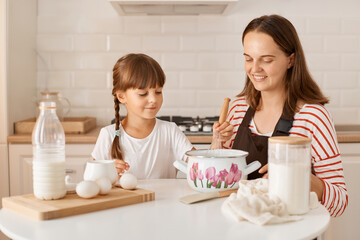 Obraz na płótnie Canvas Image of smiling dark haired female kid baking with her mother homemade pastry, sitting at table in kitchen, expressing positive emotions and happiness.