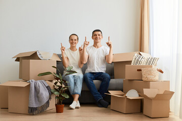 Satisfied delighted young couple sitting on sofa surrounded with carton boxes, family during relocation to a new home, looking at camera pointing fingers up.