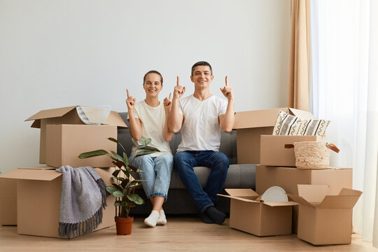 Satisfied delighted young couple sitting on sofa surrounded with carton boxes, family during relocation to a new home, looking at camera pointing fingers up. - Powered by Adobe