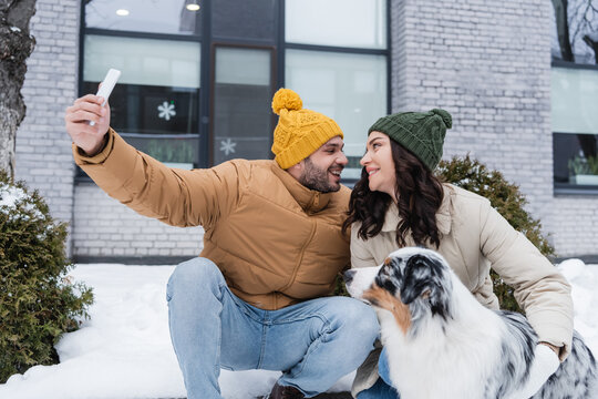 happy man in knitted hat taking selfie with cheerful girlfriend and australian shepherd dog in winter.
