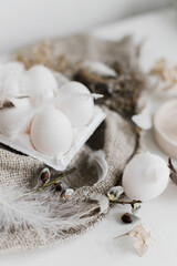 Simple rural Easter aesthetics. Natural eggs in tray, feathers, willow branches, nest on burlap on rustic white wooden table. Happy Easter. Easter rustic still life.