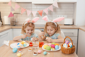 Obraz na płótnie Canvas two twin girls are preparing for the Easter holiday at the kitchen table painting Easter eggs.