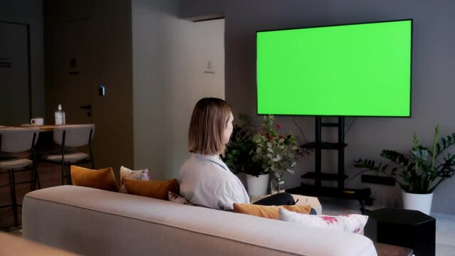 A beautiful girl is sitting on a sofa in front of a TV with a green screen. Green screen TV. Rest at home in front of the TV.