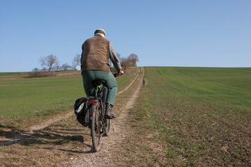 An elderly man with his bicycle rides on a field path on a beautiful spring day between the fields...