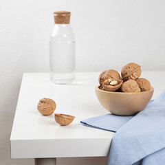 Walnuts lie in a wooden bowl standing on a white table. There is a carafe of water in the background, and a blue towel is lying next to the nuts.
